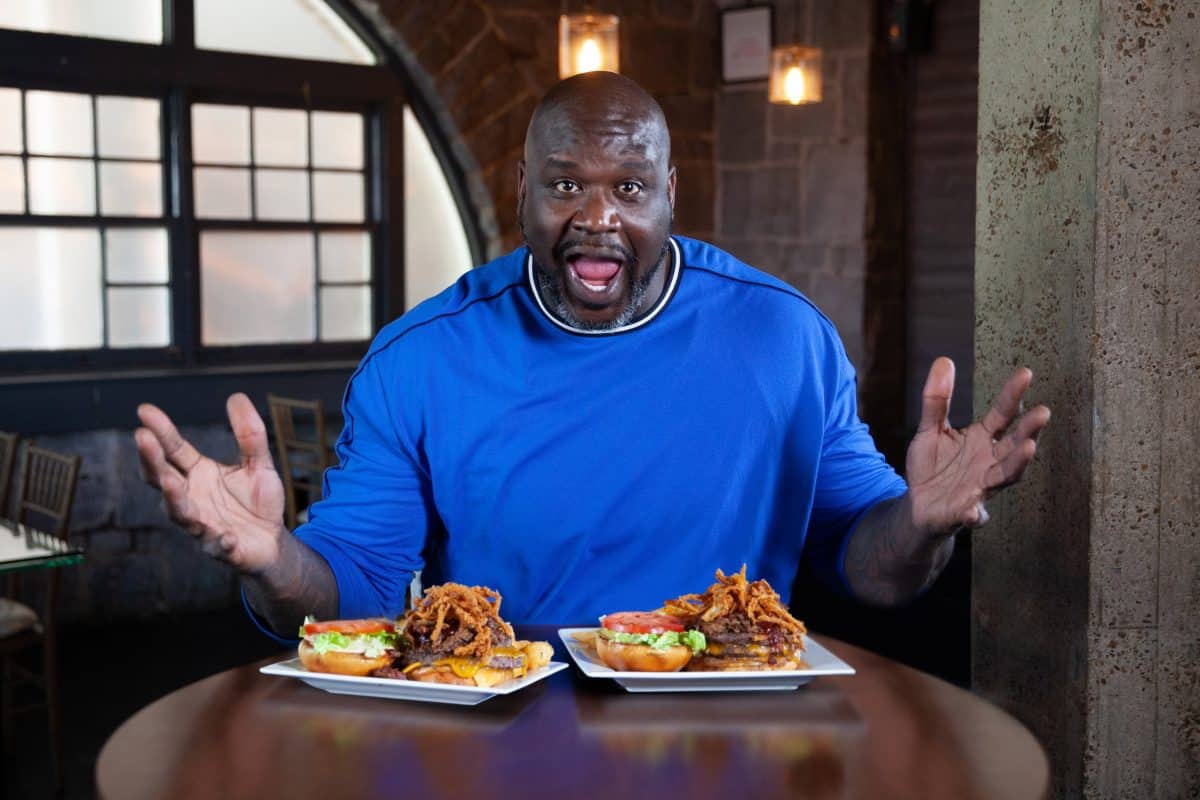 Shaq sits at a table in front of two plates with sandwiches on them.